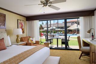 A stylish hotel room features a king bed, cozy seating, and large windows overlooking a pool area with lush greenery and lounge chairs.