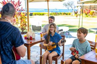 A joyful gathering features a man teaching two children to play ukulele, while another man enjoys the music in a tropical outdoor setting.