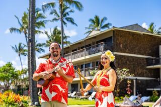 A man and woman in tropical attire play music outdoors, surrounded by palm trees and resort buildings, exuding a festive island vibe.