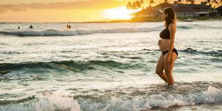 A pregnant woman stands in shallow waves at sunset, with a picturesque beach scene and gentle surf in the background.