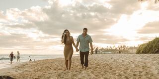 A couple walks hand in hand along a sandy beach, illuminated by a golden sunset, with people enjoying the waves in the background.
