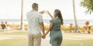 A couple holds hands, celebrating with drinks in a tropical setting, surrounded by palm trees and a serene beach backdrop.