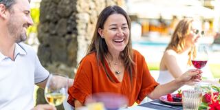 A woman in a bright orange top laughs while holding a glass of wine at an outdoor dining table, surrounded by friends and lush greenery.
