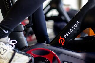 A close-up of a stationary bike with a visible Peloton logo, showcasing a rider's foot in a sneaker on the pedal.