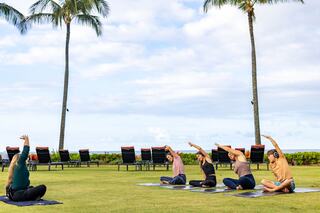 A group practices yoga on mats outdoors, surrounded by palm trees and lounge chairs, under a partly cloudy sky.
