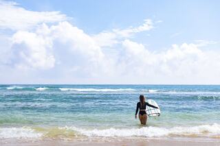 A person walks along the shore, holding a surfboard, with gentle waves and bright blue skies in the background.