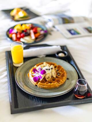 A delicious breakfast spread features a waffle topped with bananas and flowers, accompanied by fruit, orange juice, and a cozy atmosphere.