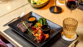 A gourmet dish featuring lobster, tender beef, and vibrant vegetables, accompanied by sauces and a glass of red wine. Elegant dining experience.
