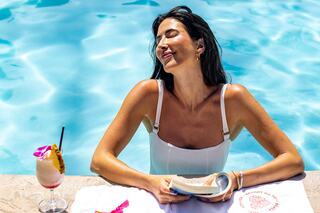 A woman relaxes poolside, savoring the sunshine with a book in hand and a tropical drink by her side, exuding a serene vibe.