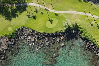 Aerial view of a lush golf course with palm trees, bordered by rocky shoreline and clear turquoise waters. A serene tropical setting.