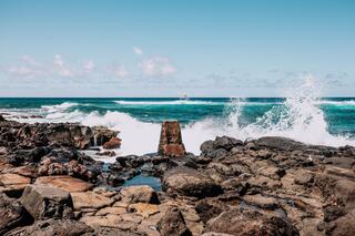 Waves crash against rocky shores under a clear blue sky, with a distant boat sailing across the sparkling ocean.