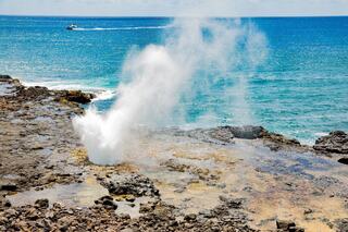 Water sprays dramatically from rocky shorelines, with a boat gliding across the vibrant blue sea in the background.