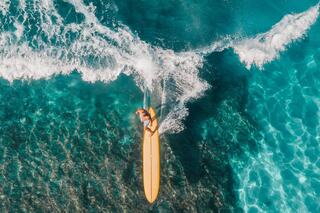 A surfer glides on a yellow board through vibrant blue waters, creating splashes against the clear ocean backdrop.