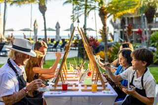 A diverse group of people enjoys an outdoor painting session, surrounded by palm trees and a beautiful ocean backdrop.