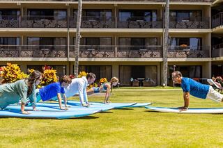 A family practices yoga or balance exercises on surfboards in a grassy area, with a hotel backdrop and tropical plants surrounding them.