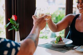 Two people toast with glasses of drink, smiling warmly. A beautiful meal and a flower centerpiece are on the table, creating a joyful atmosphere.