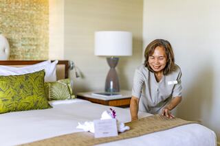 A hotel staff member smiles while arranging a neatly made bed in a well-decorated room with cozy pillows and tasteful decor.