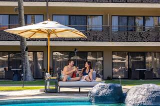 A couple relaxes by the pool under an umbrella, enjoying drinks and each other's company in a sunny, tropical setting.