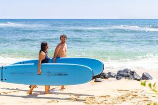 A couple walks along the beach, carrying bright blue surfboards, enjoying a sunny day by the ocean. Waves gently lap at the shore.