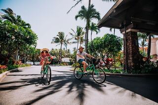 Two cyclists enjoy a sunny day, surrounded by lush tropical greenery and palm trees, creating a lively, relaxed atmosphere.
