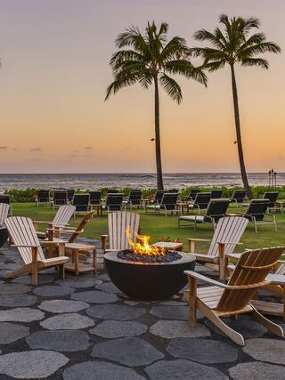 Lounge chairs surround a fire pit on a lawn with palm trees, overlooking a serene ocean at sunset. A tranquil tropical setting.