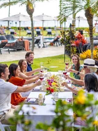 A lively outdoor gathering features a group toasting with glasses of wine, surrounded by tropical plants and a sunny poolside setting.