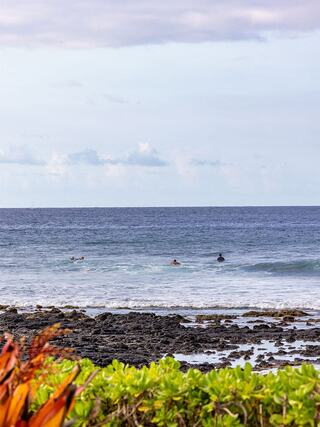Surfers in the ocean are surrounded by rocky shores and vibrant greenery under a cloudy sky, creating a serene coastal scene.
