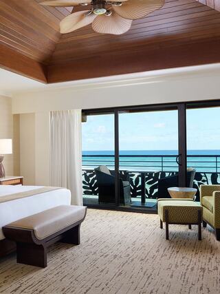 A stylish bedroom features a ceiling fan, wooden accents, a cozy bed, and a balcony with stunning ocean views. Calm and inviting atmosphere.