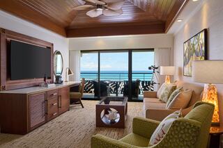 A stylish living area features a large TV, comfortable seating, and large windows offering a stunning view of the ocean and balcony.