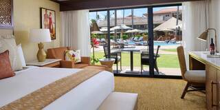 A luxurious hotel room features a king-sized bed, stylish decor, and a view of a pool area with lounge chairs and umbrellas outside.