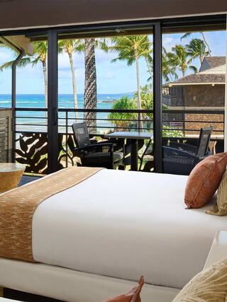 A stylish bedroom features a bed with decorative pillows, overlooking a balcony with ocean views and palm trees swaying in the breeze.