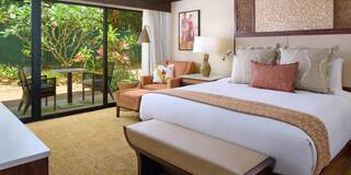 A stylish hotel room featuring a comfortable bed, earthy decor, cozy seating, and large windows opening to a lush outdoor view.