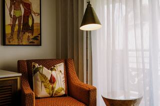 A cozy corner features an orange patterned chair with a floral pillow, a sleek table, and warm light from a stylish lamp beside sheer curtains.