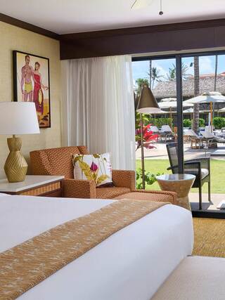 A cozy room features a patterned chair, modern lamp, and a view of a tropical patio with poolside seating and lush greenery.