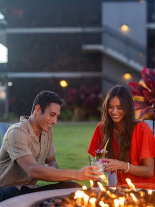 A couple toasts with drinks at a scenic table overlooking lush greenery and the ocean, enjoying a relaxed moment together.