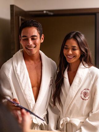 A smiling couple in cozy white robes interact warmly, exuding a relaxed, joyful vibe in an inviting setting.