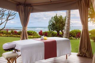 A patterned onesie with playful chicken illustrations is placed on green grass, surrounded by vibrant yellow hibiscus flowers.