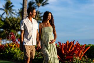A pregnant woman in a flowing green dress walks hand-in-hand with a man along a tropical pathway, surrounded by vibrant flowers and palm trees.
