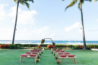 A beautiful outdoor wedding setup features rows of cushioned benches facing an ocean view, framed by palm trees and vibrant tropical plants.