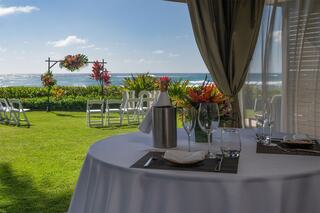A beautifully set table overlooks a lush garden and ocean, with vibrant flowers and a wedding arch in the background, creating a romantic atmosphere.