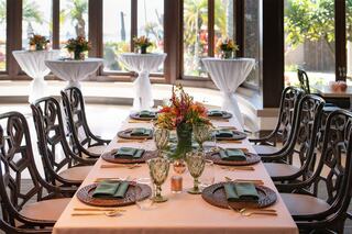 A beautifully arranged dining table with elegant tableware, surrounded by large windows, showcasing vibrant floral centerpieces and a tropical view.