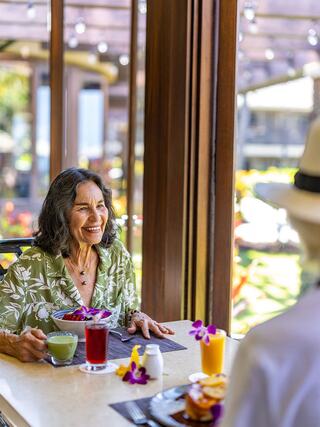 A joyful woman enjoys breakfast with colorful drinks, surrounded by bright sunlight and greenery, creating a warm and inviting atmosphere.