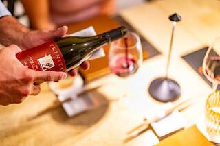 A hand pours red wine from a bottle into a glass, set against a stylish dining table with elegant decor and soft lighting.