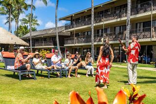 A lively outdoor performance in a tropical setting features a musician and dancer entertaining a seated audience by the poolside.