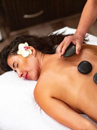 A woman relaxes during a hot stone massage, flowers in her hair, enjoying a soothing treatment in a serene environment.
