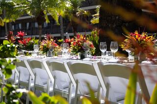 An elegant outdoor dining setup features a long table adorned with vibrant floral centerpieces and set with glasses, surrounded by lush greenery.