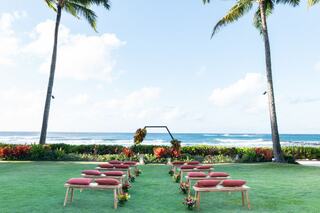 A serene outdoor wedding setup featuring rows of cushions, a floral arch, and a backdrop of palm trees and ocean waves.