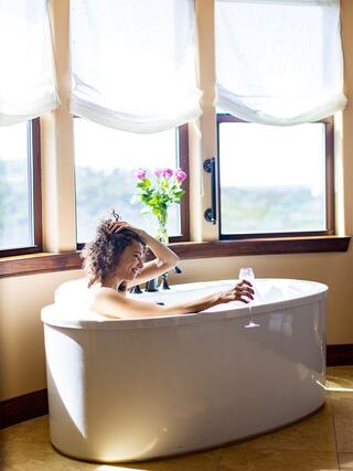 A woman relaxes in a bright bathtub, enjoying a drink, with flowers nearby and sunlight streaming through large windows.