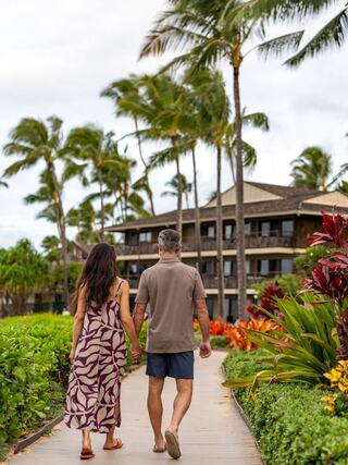A couple strolls hand in hand along a tropical path, surrounded by lush greenery and vibrant flowers, with palm trees swaying gently.
