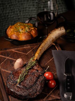 A juicy ribeye steak garnished with rosemary, accompanied by roasted garlic bread, cherry tomatoes, and a glass of red wine on a wooden board.